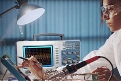 Woman working on a tablet with an oscilloscope and a soldering iron nearby